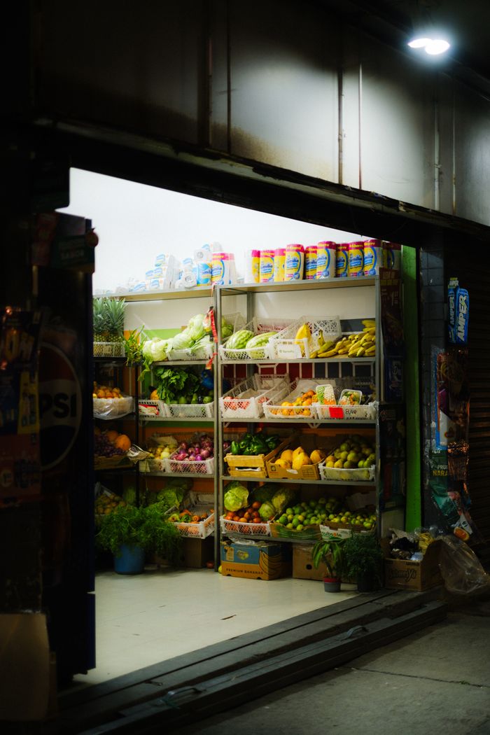 Illuminated exterior of a small grocery store in Quito showcasing fresh fruits and vegetables at night.